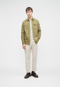 Világos olíva zöld gombos ing, két mellzsebbel, krém színű nadrággal és barna fűzős cipővel párosítva, sima háttér előtt.