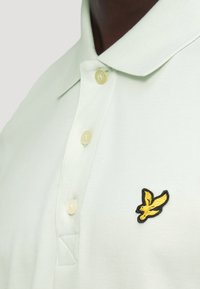 Ανοιχτοπράσινο πόλο πουκάμισο με υφή, τρία κουμπιά και λογότυπο πουλιού σε μαύρο και κίτρινο στο στήθος.