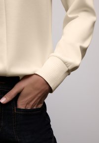 Kremowy top z długim rękawem z prążkowanymi mankietami, zestawiony z ciemnymi dżinsami. Tkanina wydaje się gładka i miękka w dotyku.