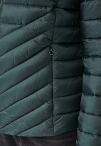 Veste verte rembourrée avec un design en chevrons matelassé, dotée d'une poche latérale zippée et d'une texture lisse.