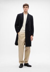 Cappotto nero doppiopetto con ampie alette, indossato sopra un maglione a coste crema e pantaloni beige, abbinato a scarpe di pelle nere.