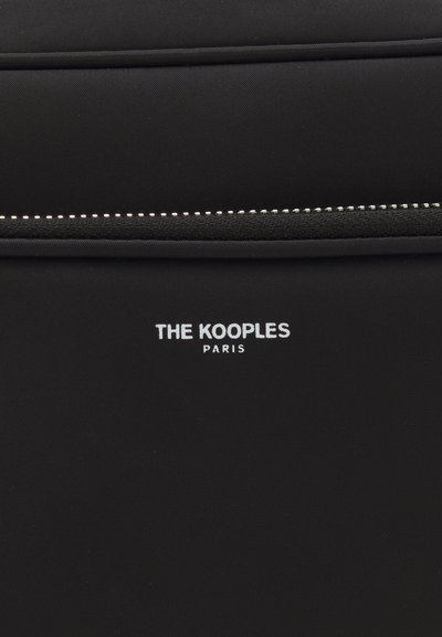 Melns auduma soma ar stilīgu dizainu, kurā baltā krāsā ir norakstīts "THE KOOPLES PARIS", un ar teksturētu rāvējslēdzēja detaļu.