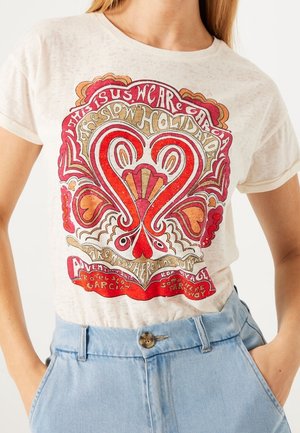 Femme portant un t-shirt graphique beige avec un motif de cœur rouge, orange et rose ainsi qu'un texte stylisé, associé à un jean taille haute bleu clair.