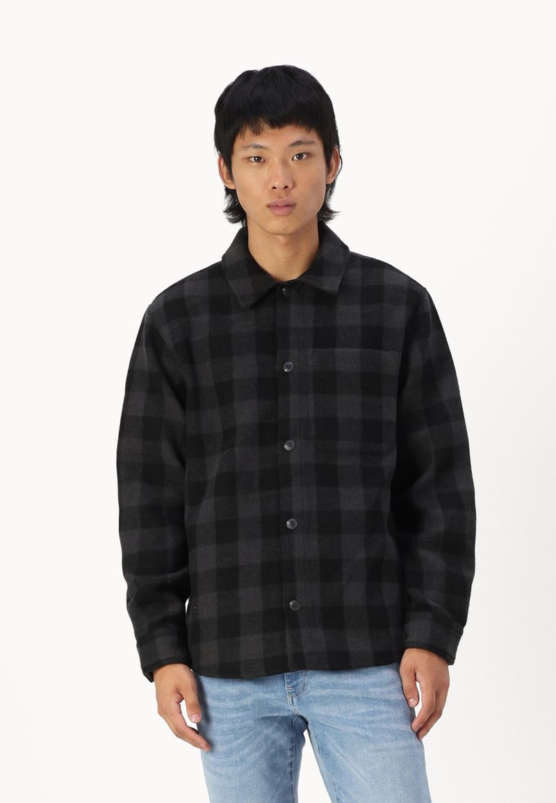 Veste en flanelle à carreaux noir et gris avec un col, fermeture à boutons et une poche poitrine. Manches longues avec une coupe décontractée.