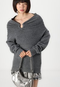 Suéter gris de punto con un escote amplio y puños acanalados, combinado con un bolso metálico plateado de forma curva y con detalle de cremallera.