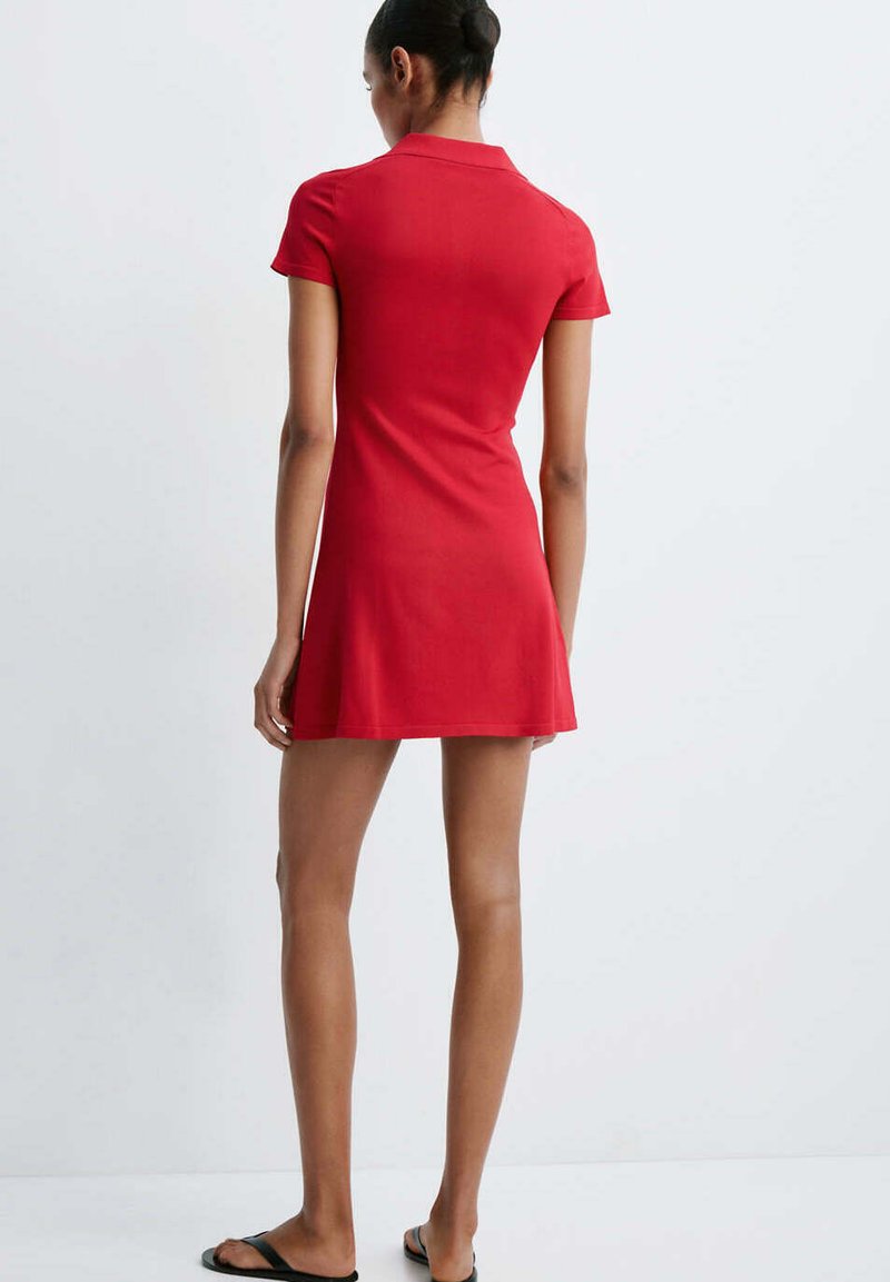 Femme avec les cheveux en chignon portant une courte robe rouge et des sandales noires, debout de dos sur un fond clair uni.