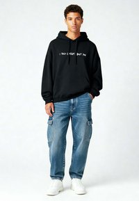 Felpa nera con cappuccio e testo bianco, abbinata a jeans cargo azzurri chiari e sneaker bianche. Stile casual con una vestibilità rilassata.