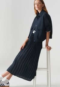 Marineblauwe cropped jacket met korte mouwen en voorkantzakken, gecombineerd met een lange geplooide marineblauwe rok. Witte sneakers en enkelkousen maken de look af.