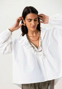 Witte blouse met gerimpelde kraag en geplooide details, gedragen met gelaagde kettingen. Lange mouwen met elastische manchetten, ontspannen pasvorm.