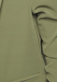 Olivgrön jacka med en jämn textur, som har en dragkedjeficka och mjukt tyg som faller. Strukturerad design med en modern silhuett.