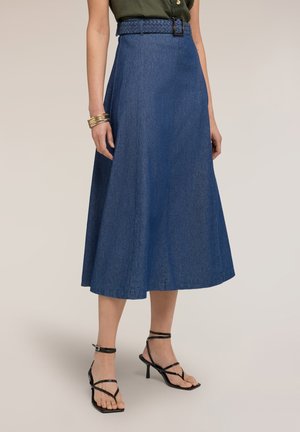 Donna che indossa una gonna midi a trapezio in denim blu a vita alta con cintura intrecciata, tacchi neri con cinturini e braccialetti dorati al polso.