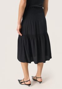 Mujer vestida con una falda midi negra con fruncidos y tacones slingback negros de punta, de pie contra un fondo claro liso, vista de espaldas.