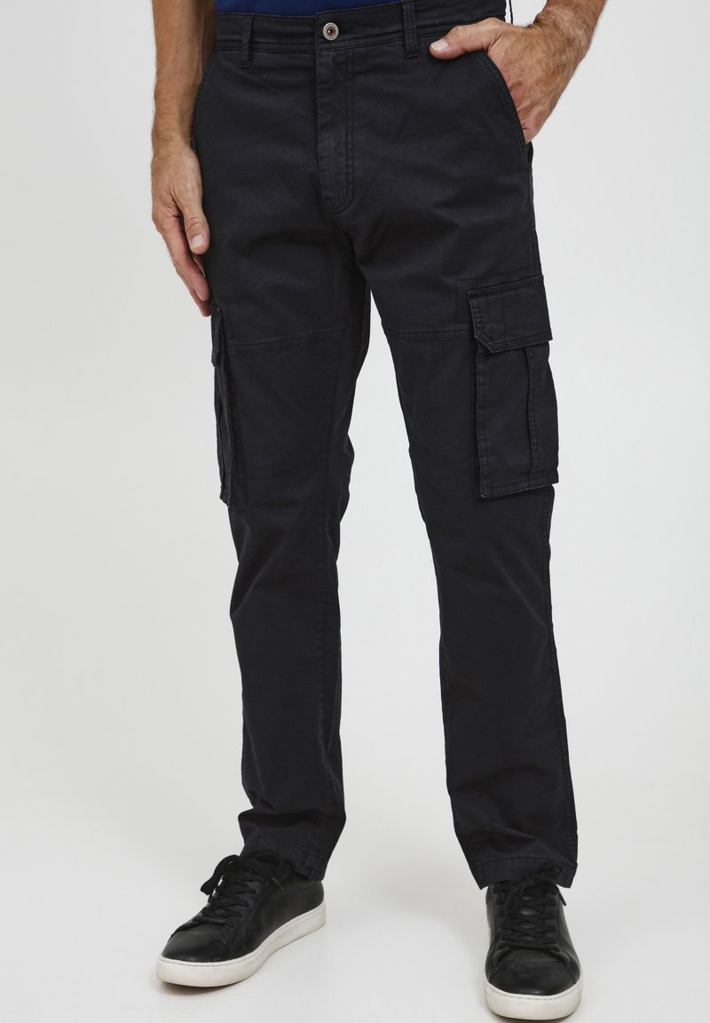 Homme portant un pantalon cargo noir avec des poches à rabat et des baskets noires, debout avec une main dans la poche, sur un fond uni.