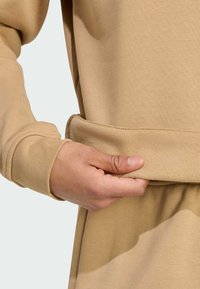 Sudadera marrón con una textura suave. Una mano agarra el dobladillo, que presenta una banda de tela de contraste ligeramente más gruesa. Se pueden ver los detalles de las costuras.