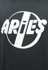 Λευκό στυλιζαρισμένο κείμενο "ARIES" με μια διαγώνια γραμμή στη μέση, περιβαλλόμενο από έναν ατελή κύκλο σε σκούρο υφασμάτινο φόντο.