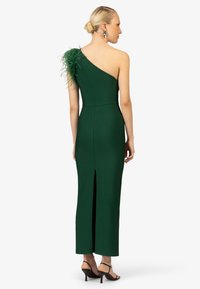 Groene een-schouder jurk met verenaccent, strakke pasvorm, zonder mouwen, rugsplit en een gladde textuur. Zwarte sandaaltjes met bandjes.