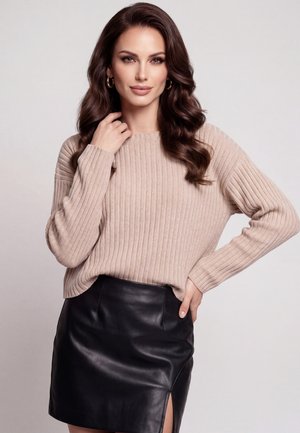Kvinde med langt bølget brunt hår iført en beige ribstrikket sweater og et sort lædernederdel med en sideslids, poserer med den ene hånd på hoften.