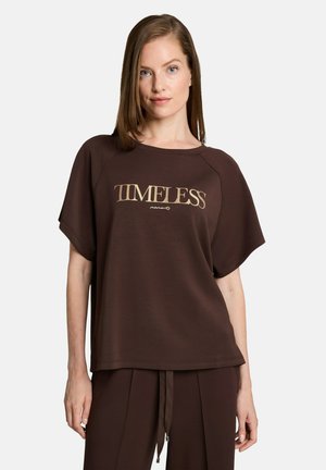 Vrouw met steil lichtbruin haar die een losse bruine blouse draagt met de tekst "TIMELESS moments" en bijpassende bruine broek op een witte achtergrond.