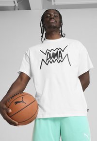 T-shirt en coton blanc avec le texte graphique noir "PUMA," associé à un short en mesh vert menthe. Le modèle tient un ballon de basket brun avec un logo.