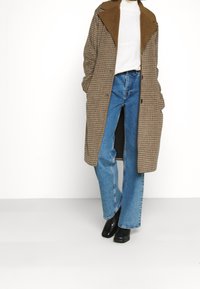 Brun rutigt kapp med bred krage, parat med ljusblå utställda jeans och svarta ankelstövlar. Har en knappknäppning och sidofickor.