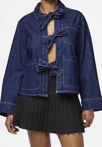 Denimjacka i mörkblått med kortad skärning, öppen framsida och rosettband. Har två fickor fram och kontrasterande sömnadsdetaljer.