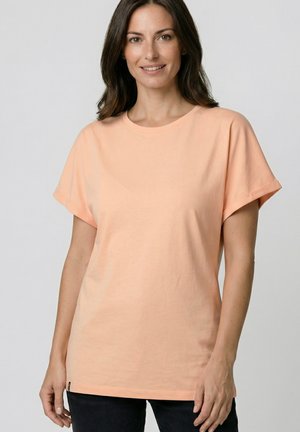 Woman with long brown hair wearing a peach short-sleeve crew neck t-shirt and black pants, standing against a plain gray background.