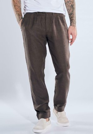 Mann trägt hochgekrempelte dunkelbraune Hose, weiße Slipper und weißes Hemd, steht mit teilweise in die Taschen gesteckten Händen.