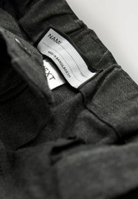 Czarny materiałowy pasek w talii z metką z informacjami "Wyprodukowano w Bangladeszu" oraz sekcjami "IMIĘ". Teksturowana powierzchnia z płaskimi szwami.