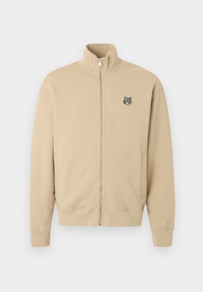 Μπεζ ζακέτα με φερμουάρ και ψηλό γιακά, με μικρό κεντημένο λογότυπο κεφαλής λύκου στο αριστερό μέρος του στήθους, διαθέτει ριμπ μανσέτες και πλαϊνές τσέπες.