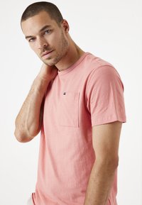 Camiseta de manga corta color coral, hecha de algodón, con un bolsillo en el pecho y escote redondo. Textura suave; diseño casual y sencillo.