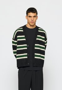 Melnā un baltā svītrainā kardigāna ar zaļiem akcentiem, ar v-veida kaklu, pogu slēgšanu un adītu tekstūru. Nēsā uz melna krekla.