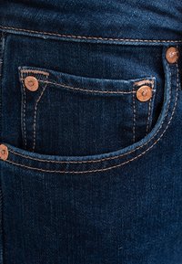 Jeans en denim bleu foncé avec des coutures visibles, comportant une petite poche avant embellie de boutons en cuivre et une texture lisse.