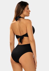 Kobieta w czarnym dwuczęściowym kostiumie kąpielowym z wiązaniem na szyi i plecach, stoi bokiem na jednolitym jasnym tle.