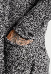 Grau gestrickter Cardigan mit einer strukturierten Musterung, ausgestattet mit einer Tasche und einer Hand mit Tattoo, die aus dem Ärmel hervortritt.