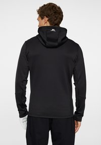 Svart huvtröja med hög krage och justerbar huva. Har en logotyp på baksidan, tillverkad av ett mjukt, elastiskt tyg. Långa ärmar.