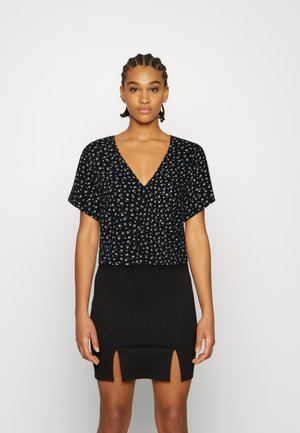 Top preto cropped com mangas curtas e decote em V, apresentando um padrão floral branco. Usado com uma saia preta com fendas laterais.