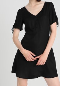 Robe noire à manches courtes avec un décolleté en V, mettant en avant des détails de boutons et des accents à nouer sur les manches. Tissu lisse ; forme de jupe évasée.