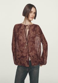 Blusa a paisley color bordeaux con maniche lunghe a palloncino. Presenta un colletto con lacci e un tessuto leggero e trasparente. Abbinata a jeans scuri.