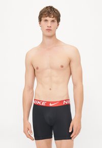 Schwarze Boxer-Shorts mit einem roten Nike-Logo-Bund. Der Stoff wirkt glatt und dehnbar, entworfen für Komfort und Unterstützung.