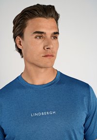 Blå, texturerad sporttröja med rund halsringning och vit "LINDBERGH"-logotyp på bröstet.