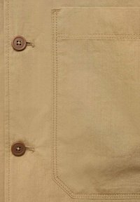 Nahaufnahme eines beigefarbenen Hemdes mit einem braunen Knopf, sichtbarer Tasche mit zartem Nähen und glattem Stoff.