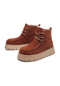 Rudi zomšiniai kulkšnies ilgio batai su paminkštu apykakle, apvaliu priekiu ir kontrastinga stambia taba platformos padais, turinčia tekstūruotą įbrėžimą sukibimui.