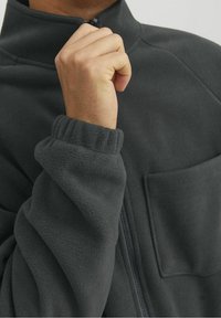 Tmavě šedá fleecová bunda s vysokým límcem, zapínáním na zip a kapsou na hrudi. Elastické manžety pro těsné přilnutí. Měkká textura.