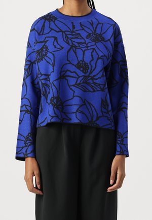 Maglione blu con grandi contorni floreali neri indossato da una persona con pantaloni neri, in piedi con le braccia rilassate ai lati.