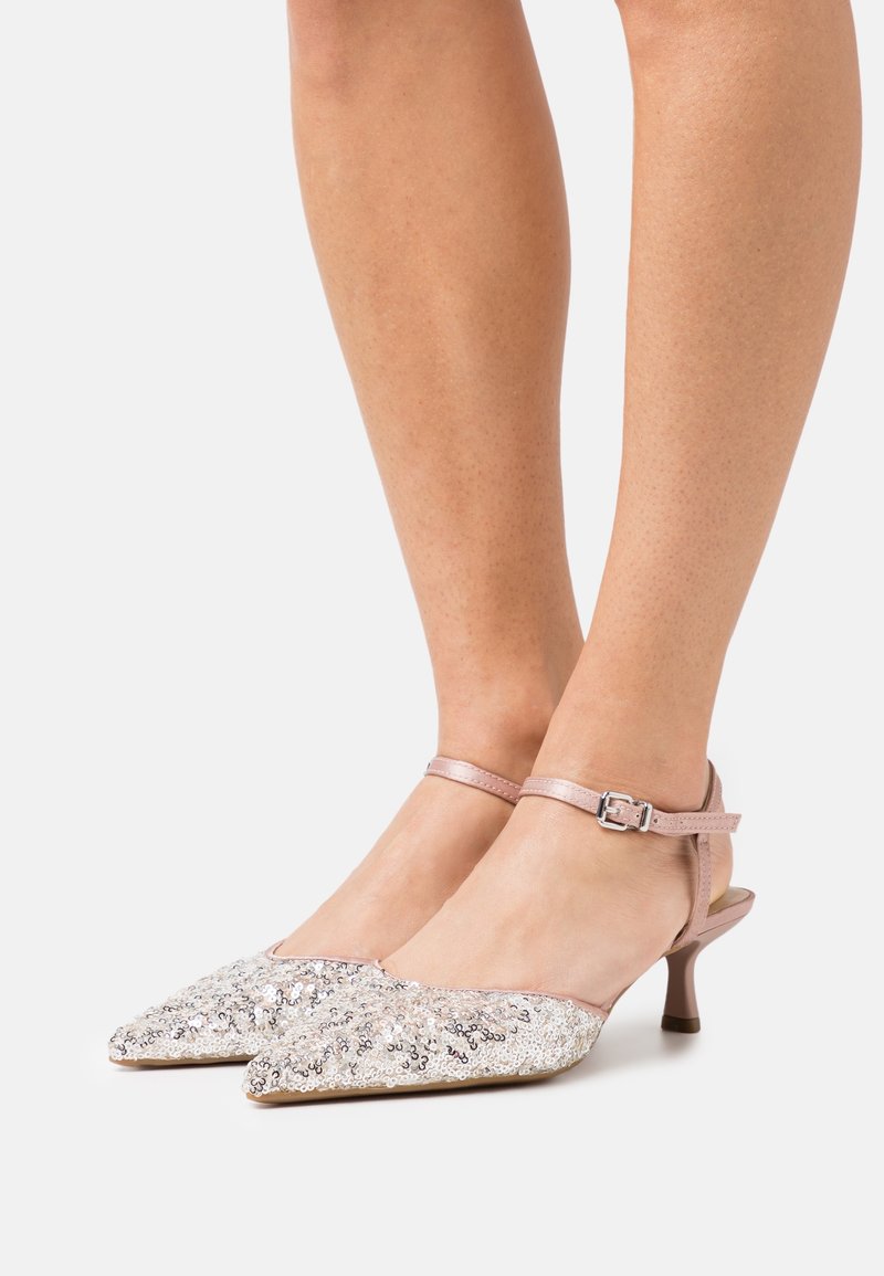 Jambes portant des chaussures roses à bout pointu, talon bas, avec des brides à la cheville et des paillettes argentées sur la zone des orteils, posées sur une surface blanche.