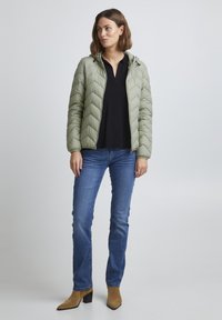 Veste matelassée vert clair avec couture en chevrons, manches longues et capuche, associée à un haut foncé, un jean bleu et des bottes à talons beiges.