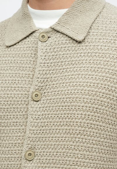 Béžová textúrovaná košeľová bunda s tkaným vzorom, vybavená špicatým golierom a dvoma gombíkmi, zdôrazňujúca pohodlný strih a jemný dizajn.