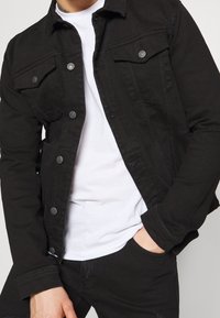 Svart denimjacka med två bröstfickor, silverfärgade knappar och klassisk krage. Bärs över en vit t-shirt med en figurnära design.