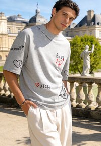 Jeune homme portant un t-shirt graphique gris et un pantalon blanc, debout en extérieur près d'une balustrade en pierre avec un bâtiment historique et une statue en arrière-plan.