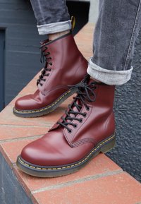 Personne portant un jean gris retroussé et des bottines en cuir bordeaux à lacets avec une couture jaune, assise sur des marches en briques à l'extérieur.